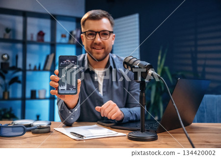 Showing graphs on the smartphone. Man with microphone in the studio, conception of podcast 134420002