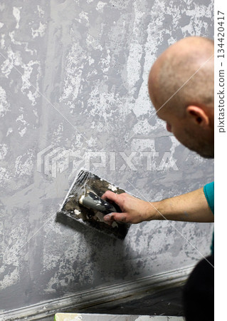 A bald man applies decorative plaster to a wall with a trowel, creating a unique, textured finish 134420417