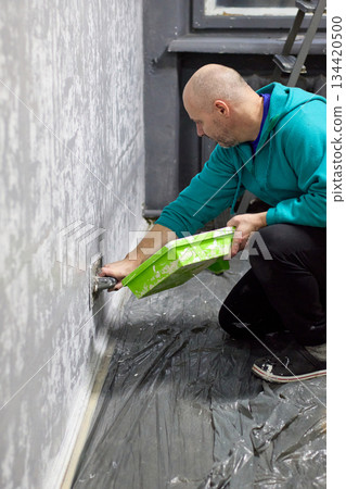 A focused bald man is carefully applying decorative plaster to a wall in a room A focused bald man is carefully applying decorative plaster to a wall in a room 134420500