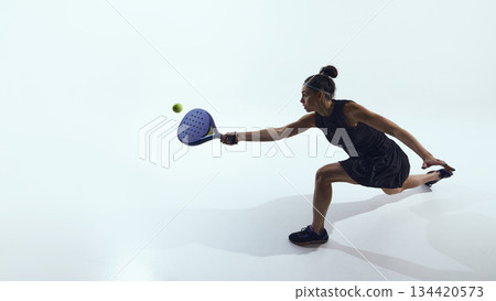 Woman lunges sideways hitting ball with padel racket in studio. Woman lunges sideways hitting ball with padel racket in studio. 134420573