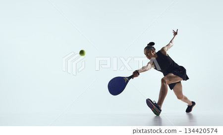 Woman stretches forward hitting ball with padel racket in studio. 134420574