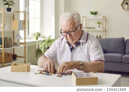 Senior man collecting wooden jigsaw puzzles game sitting at the table at home. Senior man collecting wooden jigsaw puzzles game sitting at the table at home. 134421309