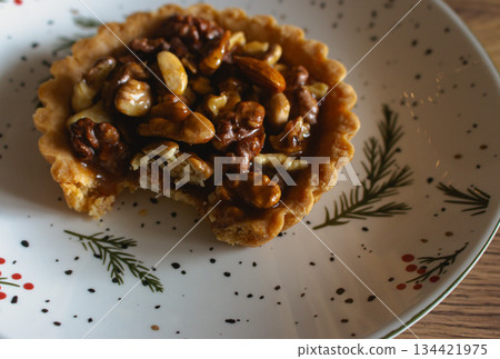 Traditional Christmas tart with nuts and salted caramel on decorated plate. Bitten tart on festive Christmas plate. Bitten hazelnut cake close up. Cinnamon cookie with caramel. Winter holiday food Traditional Christmas tart with nuts and salted caramel on decorated plate. Bitten tart on festive Christmas plate. Bitten hazelnut cake close up. Cinnamon cookie with caramel. Winter holiday food 134421975