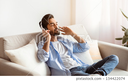 Weekend Rest. Young arab guy in wireless headphones listening audiobook at home, calm eastern man sitting with closed eyes on couch in living room, enjoying domestic leisure, free space 134422074
