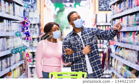 Millennial black couple in face masks shopping at grocery department of supermarket. African American family with trolley buying products at mall during coronavirus quarantine Millennial black couple in face masks shopping at grocery department of supermarket. African American family with trolley buying products at mall during coronavirus quarantine 134422107