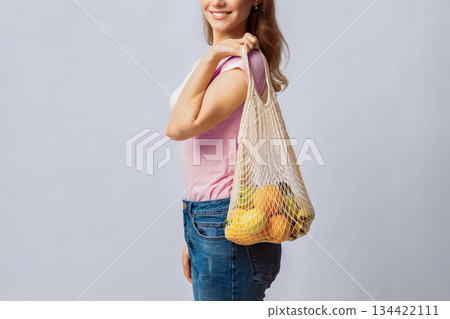 Reducing Waste. Positive Young Lady Using Reusable Eco Bag For Shopping, Unrecognizable Woman Holding Mesh With Organic Fruits And Vegetables, Standing Over Light Studio Background, Copy Space 134422111