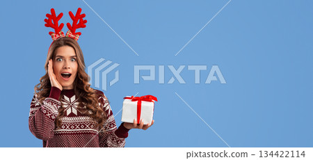 Shopping for Xmas. Surprised young lady in knitted sweater showing gift box on blue background, space for text. Panorama 134422114