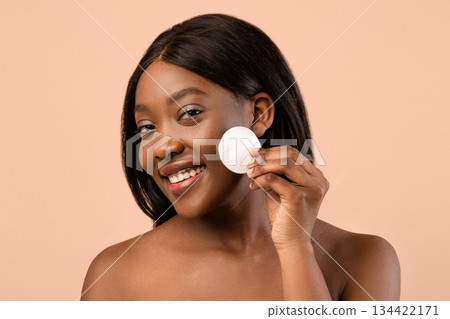 Attractive young black woman wrapped in towel cleansing face with toner, using cotton pad and facial product, closeup, beige studio background. Face care, face cleaning concept Attractive young black woman wrapped in towel cleansing face with toner, using cotton pad and facial product, closeup, beige studio background. Face care, face cleaning concept 134422171