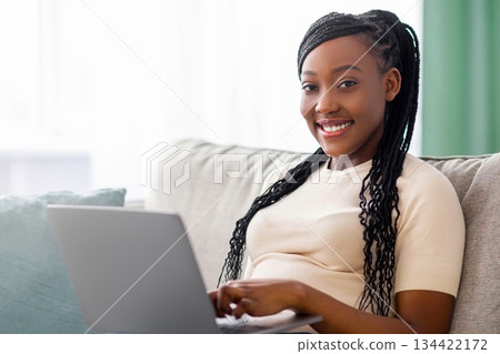 Pretty african american young lady with braids sitting on couch at home, using laptop, watching videos online, chatting with friends or reading blogs, copy space. Lifestyle during covid-19 pandemic Pretty african american young lady with braids sitting on couch at home, using laptop, watching videos online, chatting with friends or reading blogs, copy space. Lifestyle during covid-19 pandemic 134422172