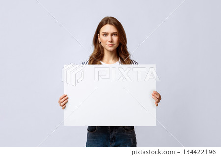 A young woman stands against a neutral background holding a large blank sign. She promotes recycling and eco-friendly initiatives with her inviting expression. A young woman stands against a neutral background holding a large blank sign. She promotes recycling and eco-friendly initiatives with her inviting expression. 134422196