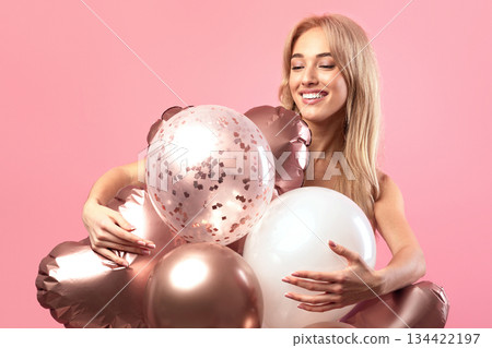 Graceful blonde birthday lady posing with colorful balloons over pink studio background. Elegant young woman expressing happiness, enjoying holiday or party, celebrating special occassion 134422197