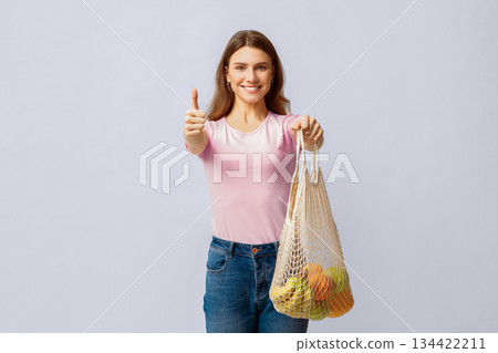 A woman stands confidently, holding a mesh bag filled with fruits while giving a thumbs-up. Her casual outfit and positive demeanor highlight her commitment to eco-friendly practices. 134422211