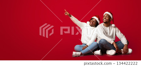 Holiday Offer. Interested Black Man And Woman In Santa Hats Looking And Pointing Away At Copy Space On Orange Background, Sitting African Couple Excited About Christmas Sale Or Promo, Panorama 134422222