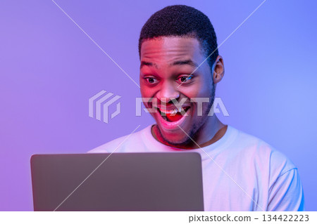 Closeup Shot Of Excited Black Guy Using Laptop Computer Under Neon Lighting, Joyful African American Man Looking At Computer Screen With Amazement, Standing Over Purple Background, Free Space 134422223