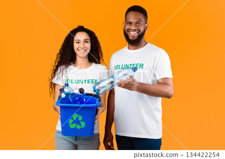 Two Cheerful Volunteers Holding Box With Recycling Symbol Full Of Wasted Plastic Bottles Standing On Yellow Background In Studio. Garbage Disposal, Sort And Recycle Your Waste Concept 134422254