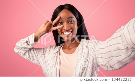 Carefree young black woman taking selfie and showing peace gesture on pink studio background. Positive African American lady smiling at camera and making photo of herself on mobile phone 134422255