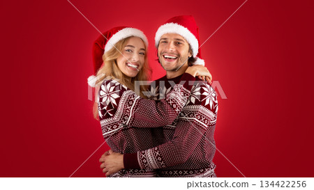 Expressing affection. Lovely young couple in Santa hats and Christmas sweaters hugging on red background 134422256