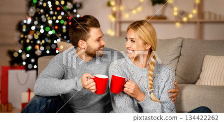Emotional Christmas couple embracing and drinking cocoa at home, copy space. Young happy man and woman sitting by festive Christmas tree and drinking tea, enjoying winter holidays together 134422262