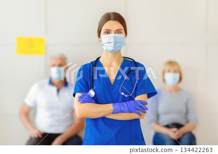 Portrait of young female doctor in uniform and face mask with patients waiting for coronavirus vaccination at clinic. Population immunization campaign, protection against covid-19 concept 134422263
