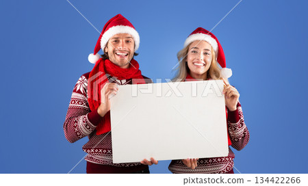 Christmas advertisement. Smiling millennial couple holding blank poster over blue background, space for design Christmas advertisement. Smiling millennial couple holding blank poster over blue background, space for design 134422266