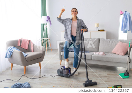 Happy young lady with vacuum cleaner having cleanup idea at messy room after party. Millennial female student hoovering dirty apartment, pointing finger up, gesturing eureka 134422267