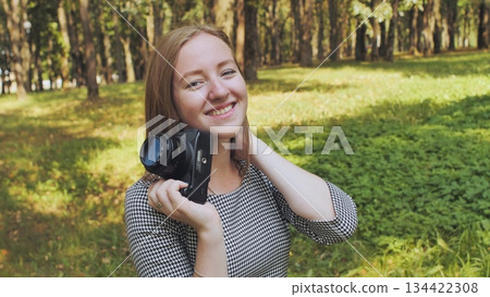 Female photographer smiling, holding camera during sunny park day, surrounded by verdant landscape and trees 134422308