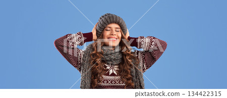 Portrait of lovely female model posing in warm knitted clothes over blue studio background, panorama. Gorgeous millennial lady wearing fashionable beanie hat, scarf and sweater with ornament Portrait of lovely female model posing in warm knitted clothes over blue studio background, panorama. Gorgeous millennial lady wearing fashionable beanie hat, scarf and sweater with ornament 134422315