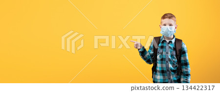 Schoolboy in protective face mask pointing at copy space over yellow studio background. Redhead schoolboy with backpack wearing face mask, using protection against coronavirus while going to school Schoolboy in protective face mask pointing at copy space over yellow studio background. Redhead schoolboy with backpack wearing face mask, using protection against coronavirus while going to school 134422317