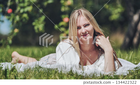 Blonde woman resting on soft blanket amid fruit trees, basking in golden sunlight during peaceful summer afternoon in verdant orchard Blonde woman resting on soft blanket amid fruit trees, basking in golden sunlight during peaceful summer afternoon in verdant orchard 134422322