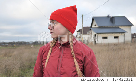 Young girl sporting braided hair, red beanie, winter jacket, gazing across rural landscape with unfinished residential construction site behind her 134422331