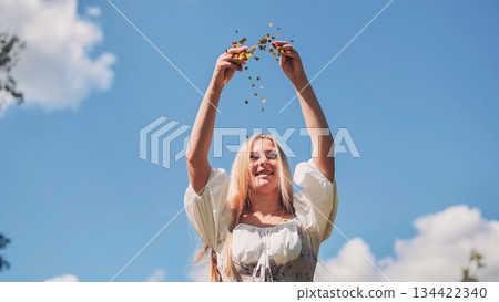 Attractive slavic woman tossing golden confetti, joyfully celebrating with vibrant blue sky backdrop Attractive slavic woman tossing golden confetti, joyfully celebrating with vibrant blue sky backdrop 134422340