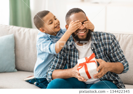 Dad's Birthday. Little Son Congratulating Black Father Giving Wrapped Gift To Him, Covering Eyes Sitting On Couch At Home. Family Holiday Celebration, Surprise Present For Daddy's B-Day Dad's Birthday. Little Son Congratulating Black Father Giving Wrapped Gift To Him, Covering Eyes Sitting On Couch At Home. Family Holiday Celebration, Surprise Present For Daddy's B-Day 134422345