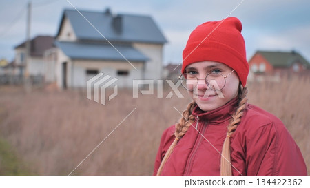 Teenage girl smiling, wearing red beanie and glasses, standing in grassy field with residential neighborhood behind Teenage girl smiling, wearing red beanie and glasses, standing in grassy field with residential neighborhood behind 134422362