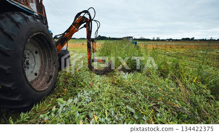 Agricultural tractor cutting grass with rotary mower, creating linear swaths on overcast farmland landscape Agricultural tractor cutting grass with rotary mower, creating linear swaths on overcast farmland landscape 134422375