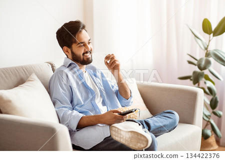 Watching TV At Home. Cheerful arab guy resting on couch and eating popcorn, smiling millennial eastern man enjoying his favorite television show, having domestic leisure, copy space Watching TV At Home. Cheerful arab guy resting on couch and eating popcorn, smiling millennial eastern man enjoying his favorite television show, having domestic leisure, copy space 134422376