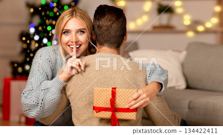 Young woman with gift box hugging her man and showing silence gesture, home interior, copy space. Loving couple celebrating winter holdiays xmas and New Year together, making surprises to each other Young woman with gift box hugging her man and showing silence gesture, home interior, copy space. Loving couple celebrating winter holdiays xmas and New Year together, making surprises to each other 134422411