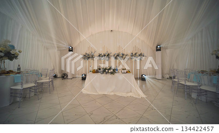 Elegant wedding table setup featuring white tablecloth, floral arrangements, and soft lighting, creating a romantic ambiance inside a spacious tent Elegant wedding table setup featuring white tablecloth, floral arrangements, and soft lighting, creating a romantic ambiance inside a spacious tent 134422457