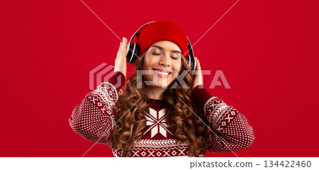 Christmas playlist. Pretty woman in warm winter clothes and headphones listening to music over red background, panorama 134422460