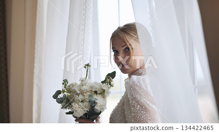 Beautiful bride holding a white bouquet and looking over her shoulder while standing near a window on her wedding day Beautiful bride holding a white bouquet and looking over her shoulder while standing near a window on her wedding day 134422465