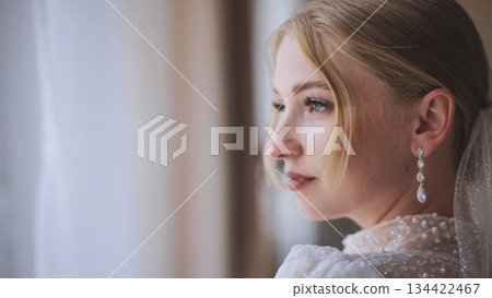 Contemplative bride with pearl earrings gazing through window on wedding morning Contemplative bride with pearl earrings gazing through window on wedding morning 134422467