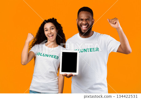 Two Multicultural Volunteers Showing Tablet With Blank Screen Gesturing Yes Standing Over Yellow Studio Background, Smiling To Camera. Activists Recommending Charity Application. Mockup Two Multicultural Volunteers Showing Tablet With Blank Screen Gesturing Yes Standing Over Yellow Studio Background, Smiling To Camera. Activists Recommending Charity Application. Mockup 134422531