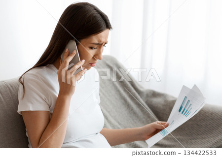 Remote Work During Pregnancy. Stressed Pregnant Woman Working With Papers And Laptop, Worried Expectant Lady Sitting On Couch In Living Room And Talking On Cellphone, Having Business Problems 134422533
