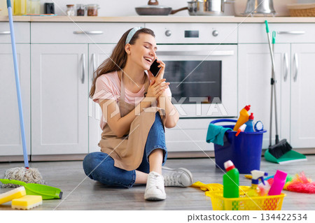Happy young housewife talking on smartphone on floor at kitchen, taking break from domestic chores to speak to her friend. Millennial lady enjoying phone conversation among cleaning supplies Happy young housewife talking on smartphone on floor at kitchen, taking break from domestic chores to speak to her friend. Millennial lady enjoying phone conversation among cleaning supplies 134422534