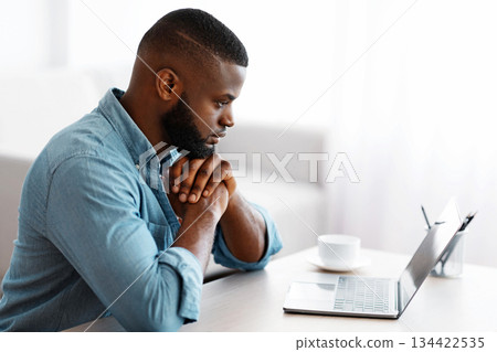 Pensive African American Man Working With Laptop At Home Office. Black Freelancer Guy Sitting At Desk And Looking At Computer Screen With Thoughtful Face Expression, Side View With Copy Space Pensive African American Man Working With Laptop At Home Office. Black Freelancer Guy Sitting At Desk And Looking At Computer Screen With Thoughtful Face Expression, Side View With Copy Space 134422535