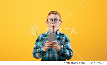 Surprised schoolboy playing game or using amazing application on smartphone over yellow studio background, copy space. Amazed ginger kid using newest mobile phone, entertainment concept Surprised schoolboy playing game or using amazing application on smartphone over yellow studio background, copy space. Amazed ginger kid using newest mobile phone, entertainment concept 134422536