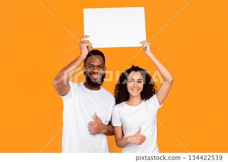 Cheerful Multiethnic Couple Holding Empty Paper Poster Above Head Gesturing Thumbs Up Posing Standing On Yellow Studio Background. Spouses Approving And Advertising Your Text. Mockup Cheerful Multiethnic Couple Holding Empty Paper Poster Above Head Gesturing Thumbs Up Posing Standing On Yellow Studio Background. Spouses Approving And Advertising Your Text. Mockup 134422539
