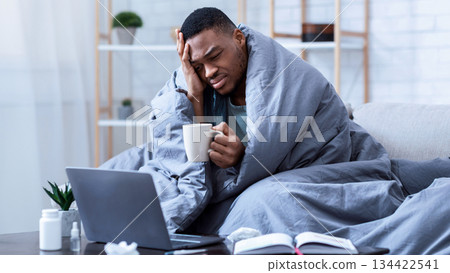 Sick African American Man Using Laptop Working While Being Ill, Having Fever And Headache Sitting On Sofa Wrapped In Blanket At Home. Guy Browsing Internet For Influenza, Cold, And Covid-19 Symptoms 134422541