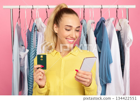 Online Payment Concept. Portrait of smiling stylish lady holding credit card and using mobile phone for shopping in internet, ordering clothes, standing with rack in the pink background 134422551
