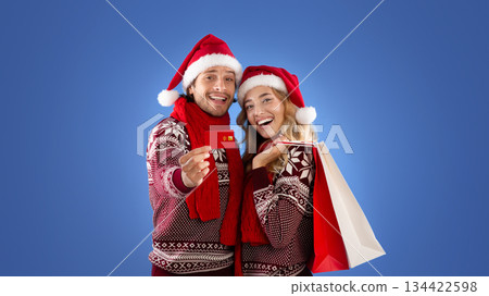Millennial couple in Santa hats showing credit card and Christmas shopping bags on blue background 134422598