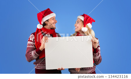 Happy young couple in Christmas outfits holding empty banner with mockup for design over blue background Happy young couple in Christmas outfits holding empty banner with mockup for design over blue background 134423116